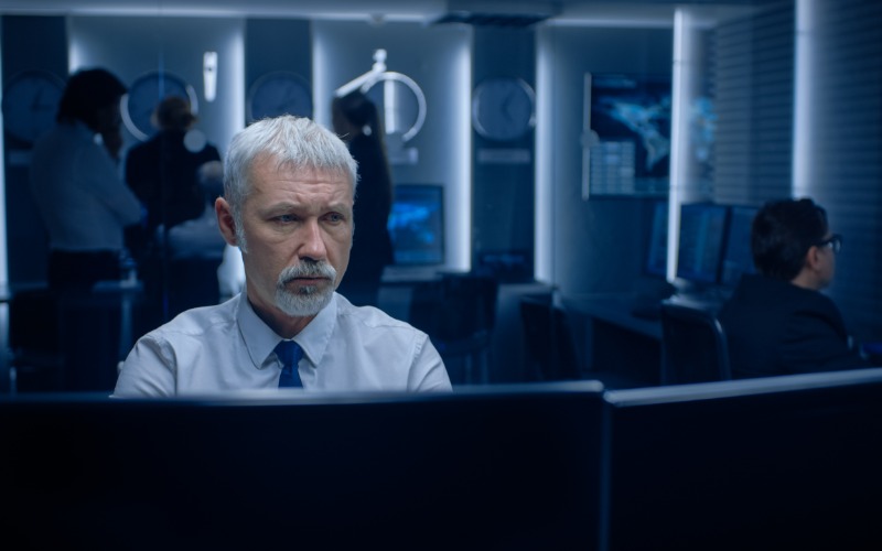 A man with a grey beard and glasses, sitting on an ergonomic black chair behind a desk with a large monitor.
